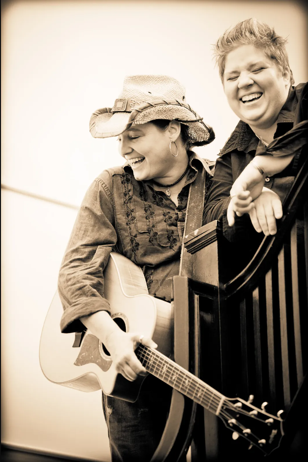 Sepia tone. Ashland Miller and Laura Cerulli lean over a stair banister, laughing. Ashland holds an acoustic guitar and wears a hat.